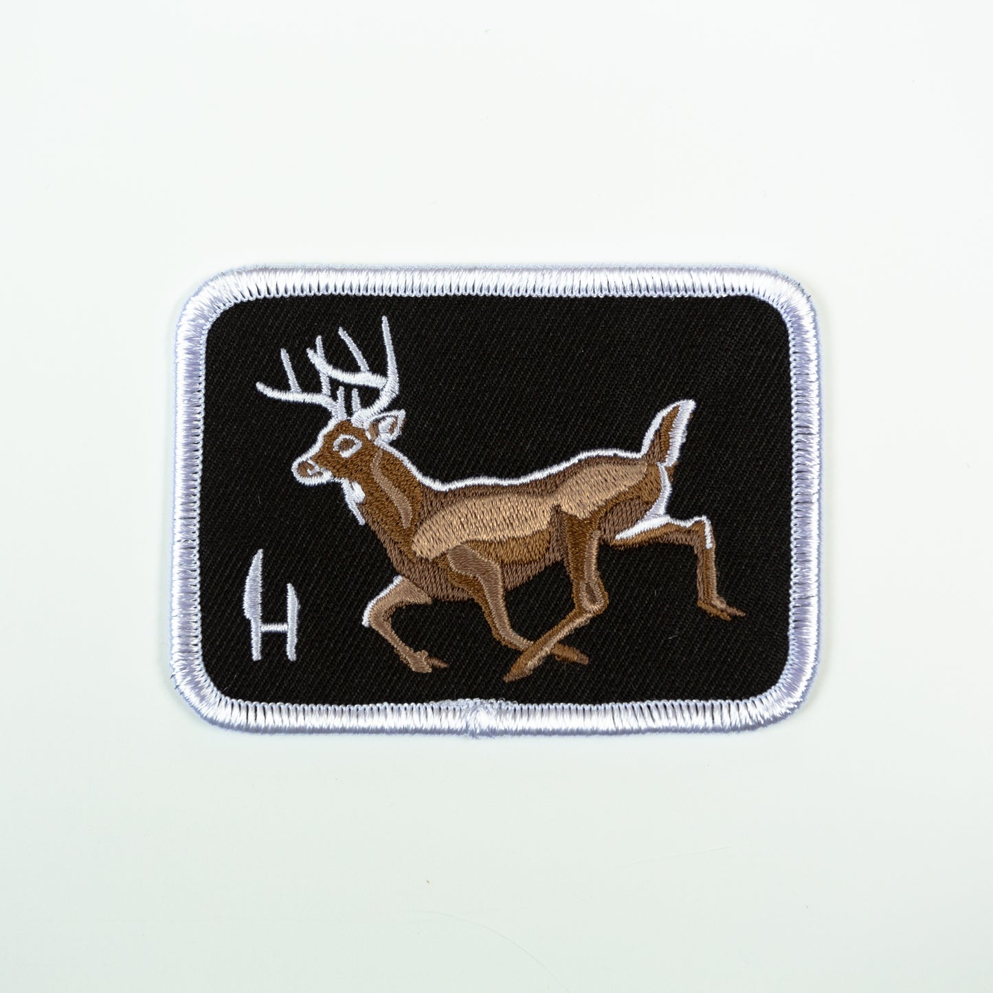 An embroidered patch of a whitetail deer buck. A black twill fabrick background with a whitetail Buck embroidered in shades of warm brown with white highlights. surrounded by a white merrowed edge and emblazened with the Hunt to eat H/knives logo