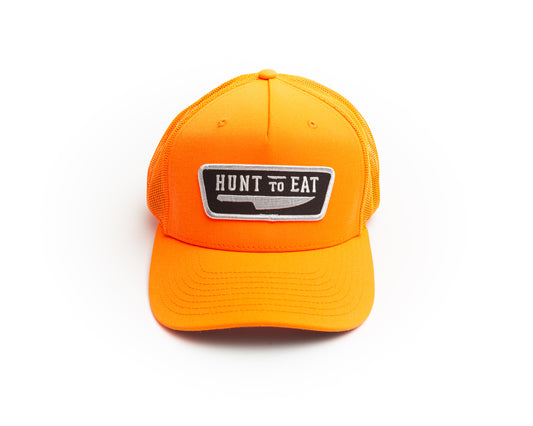 Blaze/safety Orange mesh back trucker cap.  decorated with the Hunt To Eat logo patch.  The patch contains the Hunt To Eat logo embroidered in white stitching on a black twill background surrounded by a white thread merrowed edge border. safety orange for staying visible while hunting during gun seasons. 