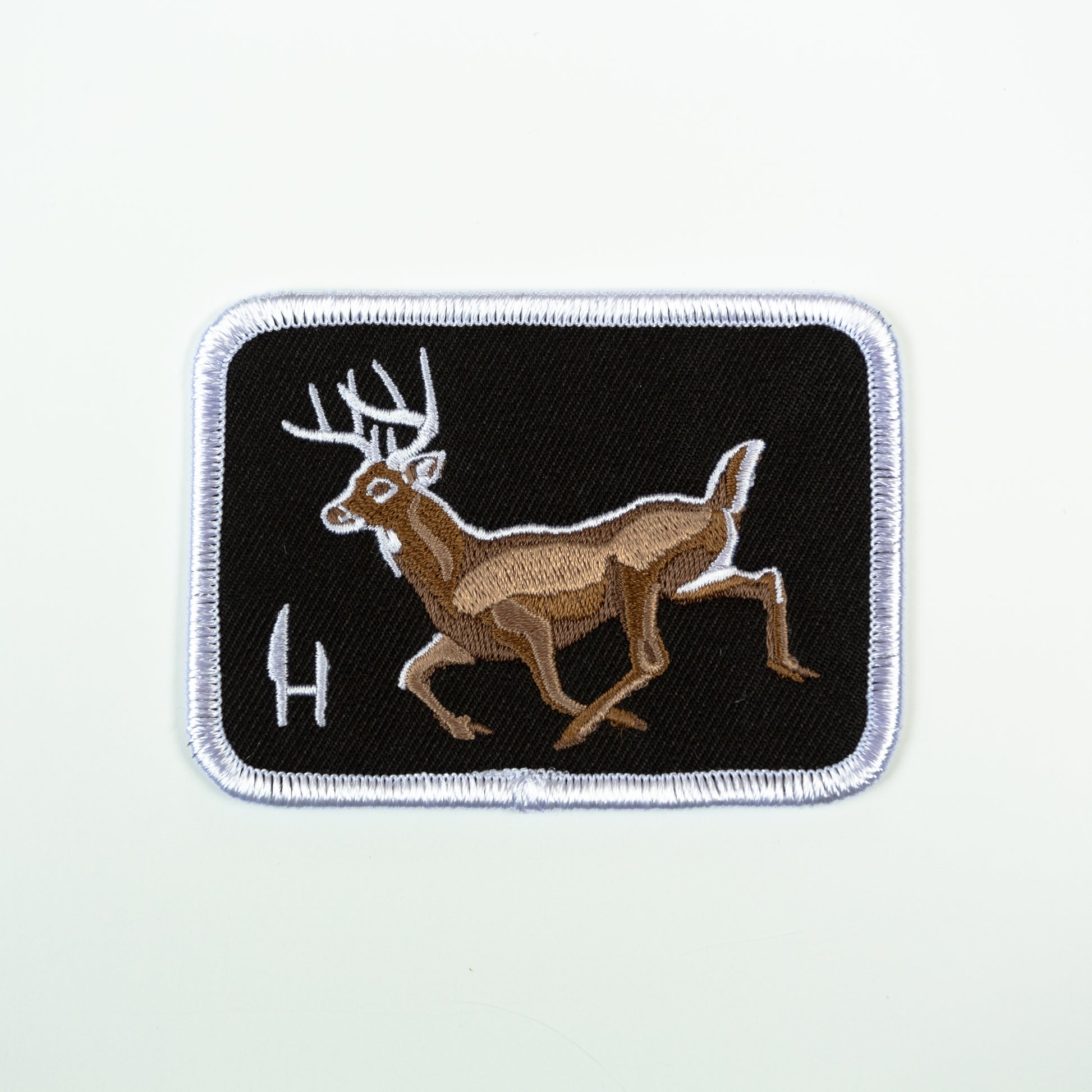  An embroidered patch of a whitetail deer buck.  A black twill fabrick background with a whitetail Buck embroidered in shades of warm brown with white highlights.  surrounded by a white merrowed edge and emblazened with the Hunt to eat H/knives logo